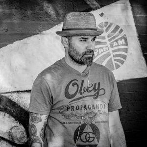 Grayscale photo of artist Adrian "Viajero" Roman. He wears a hat and looks away from the camera with a neutral expression.