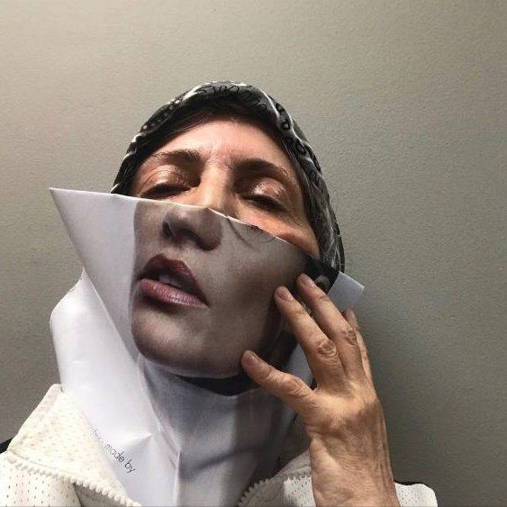 A close up photo of artist Rodríguez Calero holding a folded photo of the lower half of a woman's face over the lower half of her own face, obscuring it. She looks at the camera with nearly closed eyes.