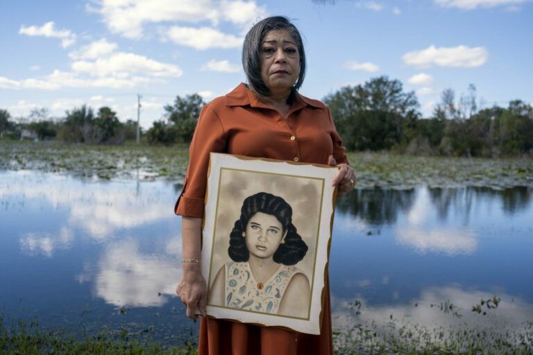 A woman dressed well solemnly stands holding a painted portrait of another woman. She stands in front of a body of water.