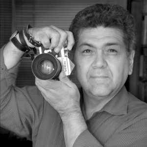 A grayscale self portrait of artist George Malave. He holds the camera next to his face and smiles slightly at the viewer.