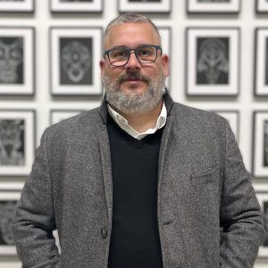 A photo of artist Juan Alberto Negroni in an art gallery. He wears glasses and smiles lightly at the camera.