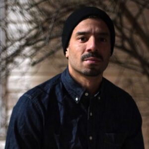 A photo of artist Christopher López. He stands half in the shadow and looks at the camera with a neutral expression.