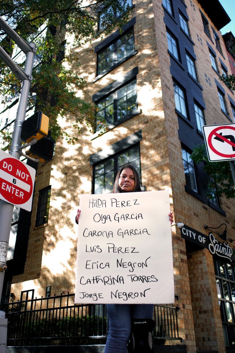 Woman standing in the street with a sign that reads "Hilda Perez, Olga Garcia, Carona Garcia, Luis Perez, Erica Negron, Cathrina Torres, Jorge Negron"