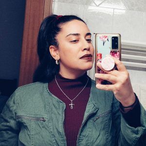 Artist Amanda Raquel Dorval taking a selfie in a mirror. She looks at her phone with a neutral expression.