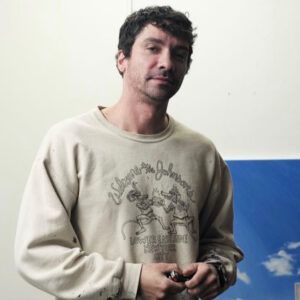 A headshot of artist André Marcel Pagán. He stands in front of his artwork with a neutral expression.