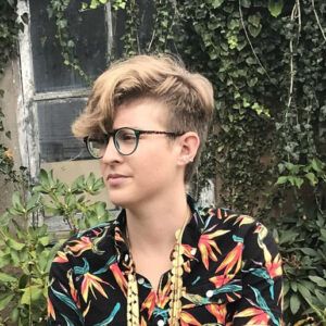 Artist Andrea Pérez Bessin stands in front of a plant covered building. They wear glasses and look away from the camera with a neutral expression.