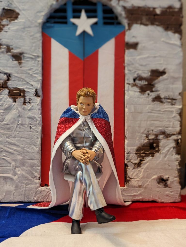 3D printed Walter Mercado statue wearing a silver suit and a Puerto Rican Flag cape. he sits with this legs crossed and his hands folded. Behind the statue there is a doorway with the Puerto Rican flag as the pattern for the double doors.