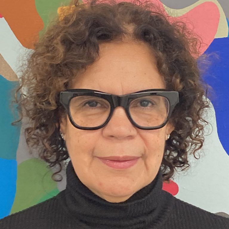 A close up photo of artist Candida Alvarez standing in front of her artwork. She wears glasses and smiles at the camera.