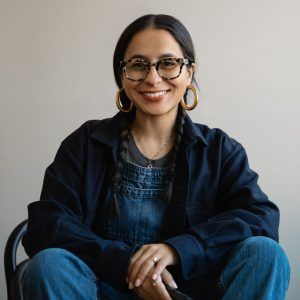 A photo of artist Daniela Spector. She sits in front of a blank wall and grins at the camera.