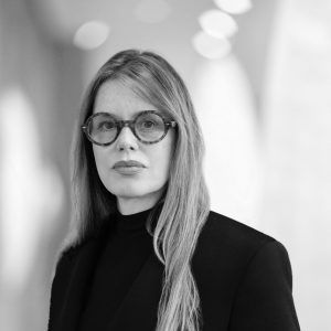 A grayscale close up photo of artist Gisela Colón in front of a blurred background. She wears glasses and looks at the camera with a neutral expression.