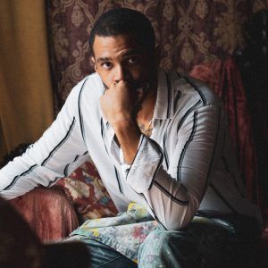 A headshot of artist José Angel Báez Christopher sitting half in shadow. His head is propped on his hand, obscuring his mouth and he looks into the camera with a neutral expression.