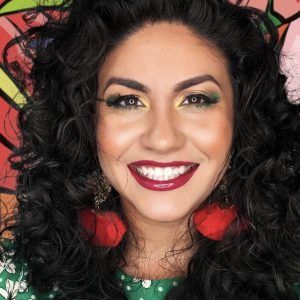 A close up photo of artist Sketzii in front of her artwork. She grins at the camera.