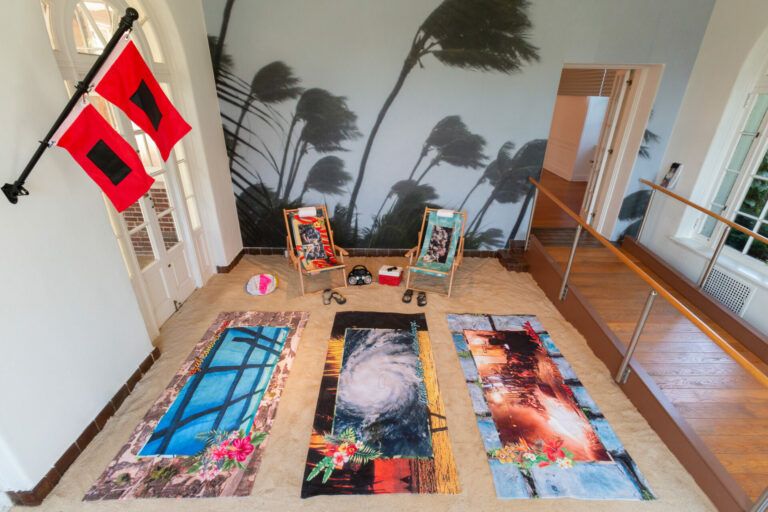 Beach towels in a room staged like a beach. Each towel has a different print which includes a border and a floral accent. There are beach chairs and accessories.
