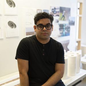 A photo of artist Kevin Quiles Bonilla. They are seated at a desk in their studio. He wears glasses and looks into the camera with a neutral expression.