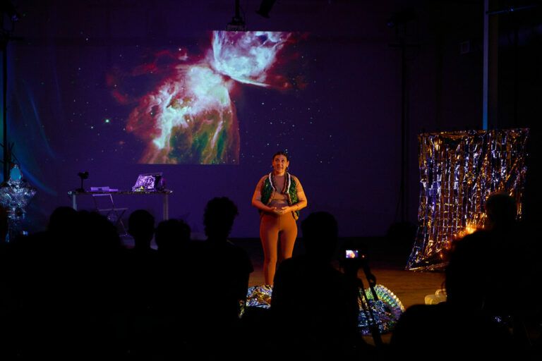 A film still of a person standing on stage with a shiny vest. Surrounding the performer there are images of space and reflective objects. There is a table in the background with a lamp and supplies.