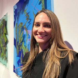Artist Livia Ortiz Ríos stands in a gallery displaying her artwork behind her. She grins at the camera.
