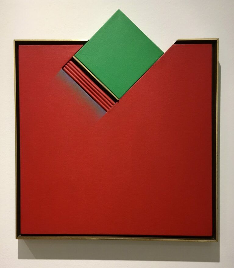 An almost square red painting. The top side of the square is interrupted by a green square jutting out the frame. Underneath the green square, there are small red ridges and a blue gradient.