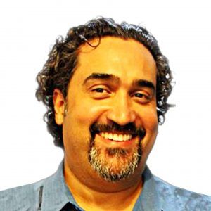 A close up photo of artist Luís A. Pagán. Their head is turned slightly and he grins at the camera.