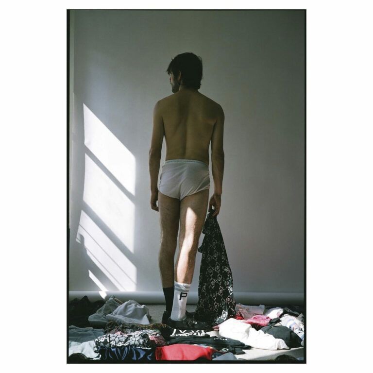 A masculine person stands in a pile of clothes wearing nothing but underwear, socks, and sneakers. Their back is to the viewer and they hold a patterned piece of clothing.