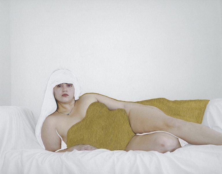 A figure lounges on white fabric. They are enamel painted white over their hair and their torso is enamel painted in a gold color.