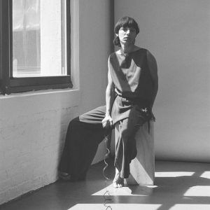 A grayscale portrait of artist Mara Corsino seated. She holds a cord and looks at the camera with a neutral expression.