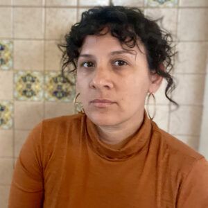 A photo of artist Mireya Lucio. They are sitting in front of a tiled wall. She looks at the camera with a neutral expression.