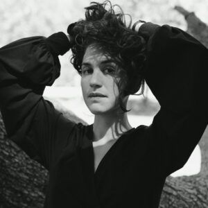 A close up grayscale photo of artist Natalia Lassalle-Morillo standing in front of a tree with her hands behind her head. She looks at the camera with a neutral expression.