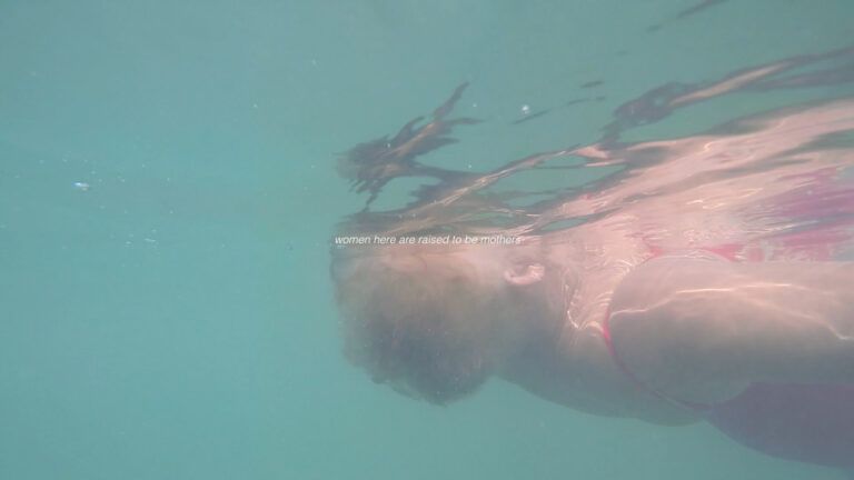 Film still of a woman floating face up in a body of water. The perspective is such that the viewer is under water and the woman's face is obscured above the water. The text reads "women here are raised to be mothers."