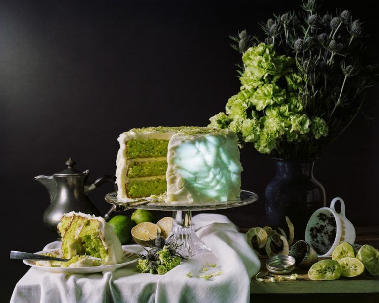 A pale green still life with cake, citrus fruits, flowers, and tea. A muscular body is projected onto the cake icing.