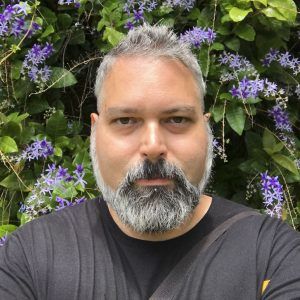 A close up photo of artist Néstor Pérez-Molière in front of a flowers. He looks at the camera with a neutral expression.