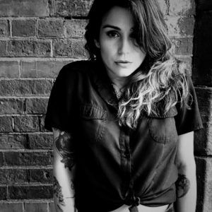 A grayscale photo of artist Nicole "Niko" Bueso standing in front of a brick wall. She looks at the camera with a neutral, open expression.