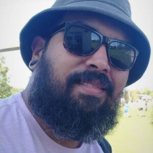 A close up photo of artist Roberto Marquez. He wears a hat and sunglasses and grins lightly at the camera.