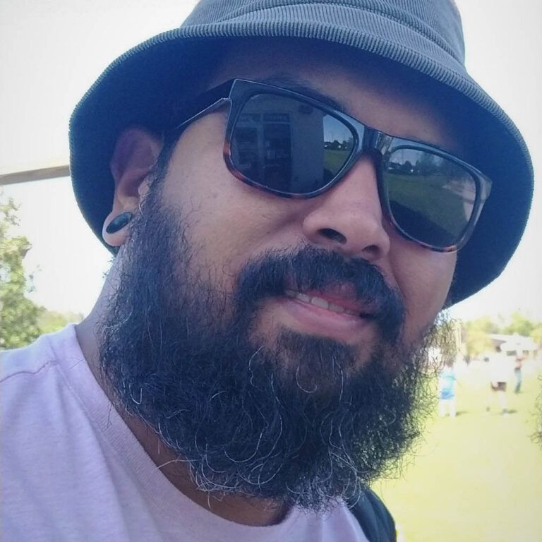 A close up photo of artist Roberto Marquez. He wears a hat and sunglasses and grins lightly at the camera.