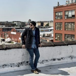 A photo of artist Sebastian Vallejo standing on a rooftop with other building behind him. He wears a hat and sunglasses and faces away from the camera.