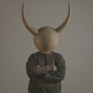 A headshot of artist Diego Antonio Espaillat. He wears a piece of his artwork, a mask with large horns.