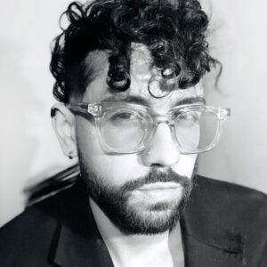 A grayscale close up photo of artist Lionel Cruet. They wear glasses and look at the camera with a neutral expression.