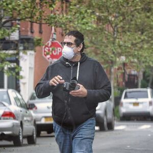 A photo of artist David Gonzalez at work. He wears glasses and an n95 respirator as he walks down the street holding a camera.