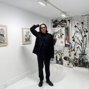Artist Fernando Pintado stands with a hand on his head in a gallery displaying his work. He wears glasses and looks at the camera with a neutral expression.