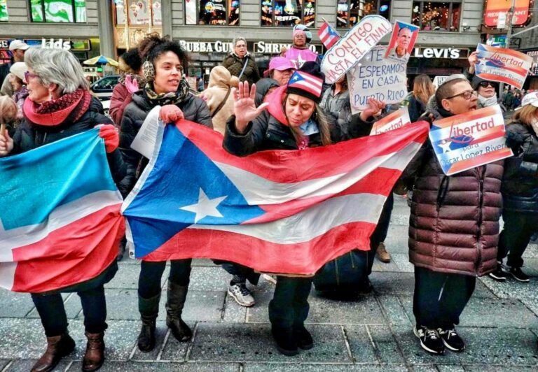 Women at a rally carrying signs about liberty and Oscar Lopez. They hold up Puerto Rican flags.