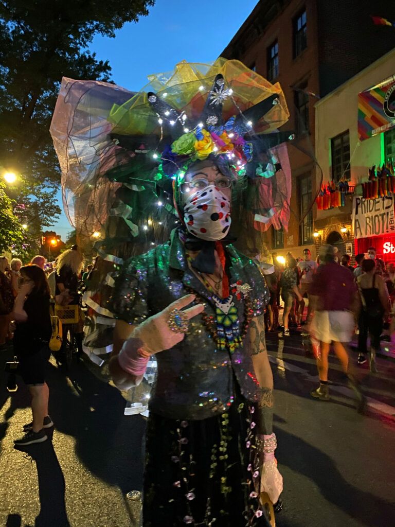 Pride march during COVID. The central figure wears a flower headband with tuille and lights on their head. They have a mask covering their nose and mouth. They are wearing sparkly jewelry and pearls.