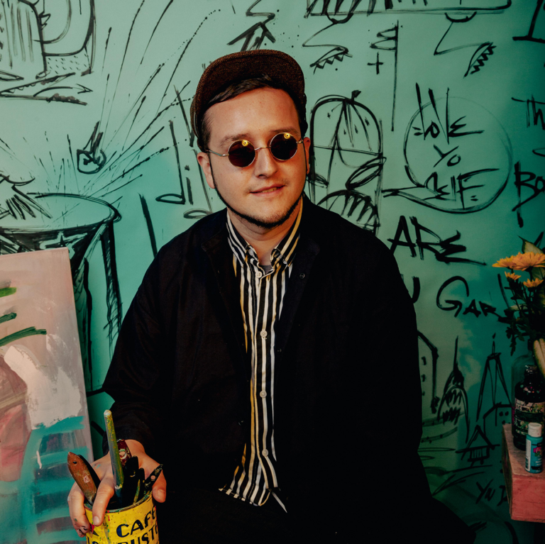 Headshot of artist Adrian Bermeo, He sits in front of his artwork smiling while wearing a hat and sunglasses.