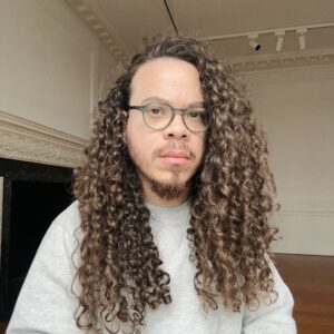 Artist Carlos Wilfredo Encarnación stands in an empty gallery. He wears glasses and looks at the camera with a neutral expression.