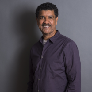 A headshot of artist Pepón Osorio. He stands in front of a grey backdrop and grins at the camera.