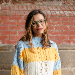 Artist Cindy Lozito stands in front of a brick wall. She wears glasses and smiles at the camera.