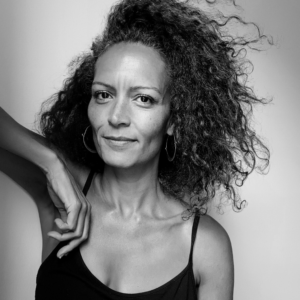 A grayscale photo of artist Wanda Benvenutti with her arm propped against something out of frame. She smiles into the camera.