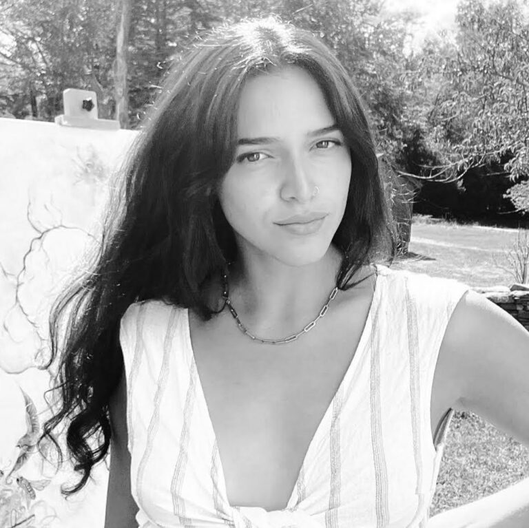 A grayscale photo of artist Kayleenda standing in front of an easel. She smiles lightly at the camera.