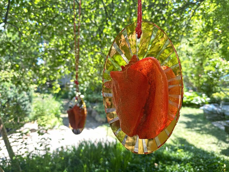Conch shell resin molds encased in a mirrored circle. The objects hang from a tree.