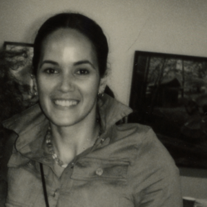 A grayscale photo of artist Melanie Gonzalez. She faces the camera and grins.