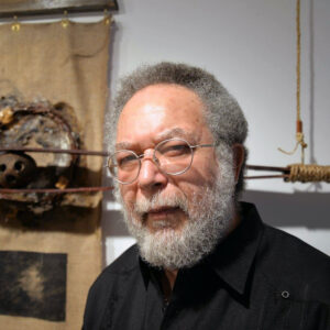 Artist Luis Stephenberg Alers stands in front of one of his sculptures. He wears glasses and looks at the camera with a neutral expression.