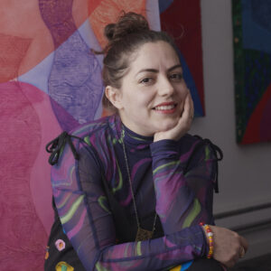 Artist Nora Maité Nieves sits in front of her artwork. Her head is propped up on her hand and she grins at the camera.